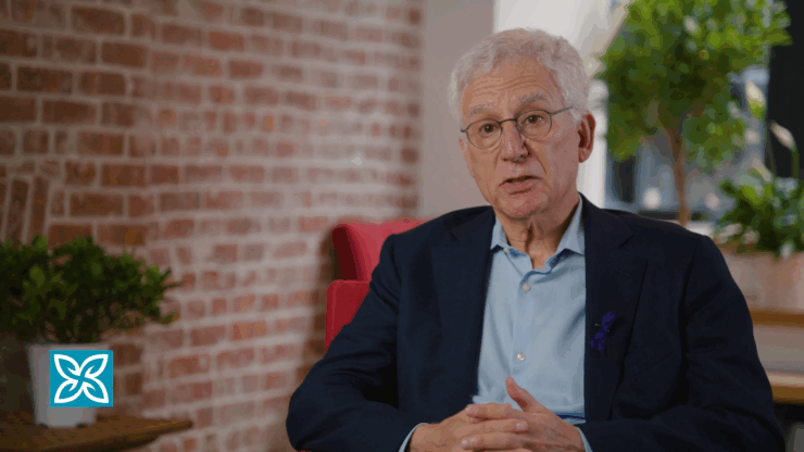 Elderly man with gray hair and glasses speaks while seated. He's wearing a suit with a blue shirt.